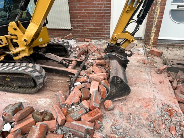 Compacte minigraver sorteert bakstenen en sloopafval naast een woning