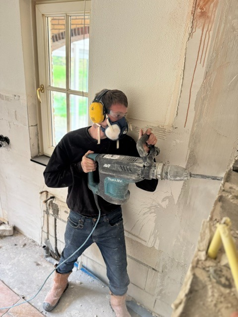 Vakman met gehoorbescherming en stofmasker breekt met een breekhamer een binnenwand open